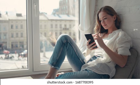 Close up pretty young woman sitting on a windowsill at home and texting on her phone communication female looking message cellphone cheerful smile use internet modern smartphone slow motion