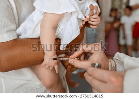 Close up of priest hand using small metal brush for chrismation on infant palm held by parent during christening ritual in church religious tradition of anointing baby with oil and faith