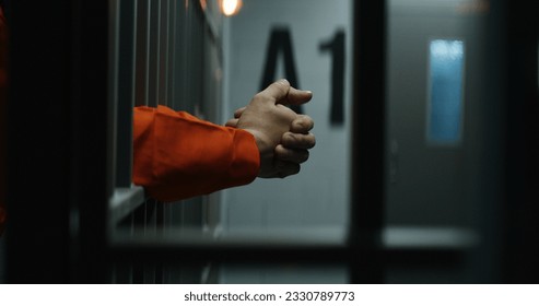 Close up of prisoner in orange uniform holding metal bars, standing in prison cell. Guilty criminal or killer serves imprisonment term for crime. Inmate in jail or detention center. Justice system.