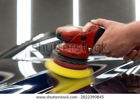 Close up of a professional car detailing process. Mans hand holds a red electric orbital dual action polishing machine with a yellow foam pad buffer tool to polish the dark blue paint gloss surface of a luxury vehicle hood in a garage workshop.