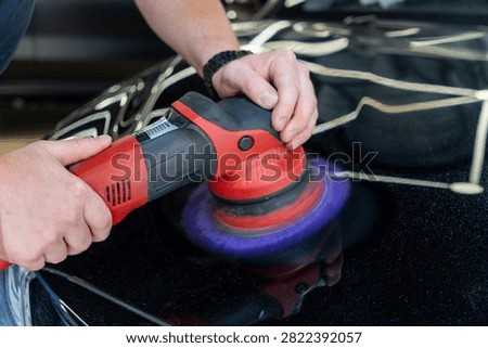 Close up of a professional car detailing process. Mans hands hold an electric red orbital dual action polisher machine with a purple foam pad buffer tool to polish a black auto paint hood surface in a garage vehicle maintenance workshop shop.