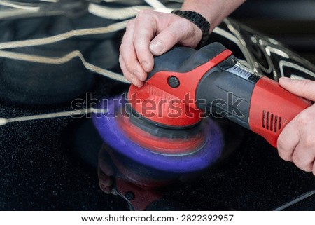 Close up of a professional car detailing process. Mans hands hold a red electric orbital dual action polisher machine with a purple microfiber wool pad buffer tool to polish a black auto paint hood surface in a garage vehicle maintenance workshop.
