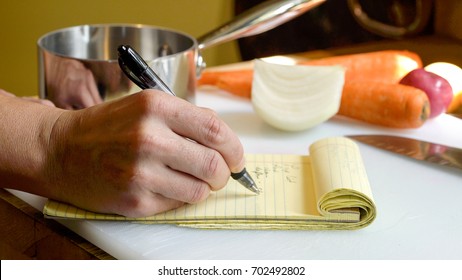 Close Up Of A Professional Chef's Hand, Writing A Mise En Place Food Prep List On A Yellow Notepad To Stay Organized In Preparing A Meal.  Vegetables And Kitchen Tools In The Background. 