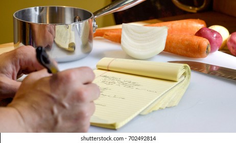 Close Up Of A Professional Chef's Hand, Writing A Mise En Place Food Prep List On A Yellow Notepad To Stay Organized In Preparing A Meal.  Vegetables And Kitchen Tools In The Background. 