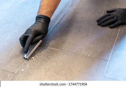 A close up of a professional cleaner cleaning grout with a brush blade and foamy soap on a gray tiled bathroom floor