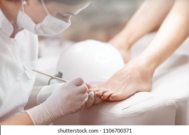 Close up of professional cleaning cuticle around nails on feet, with wooden stick. Female specialist in mask on face, making pedicure for client with uv lamp near. Beauty salon and service.