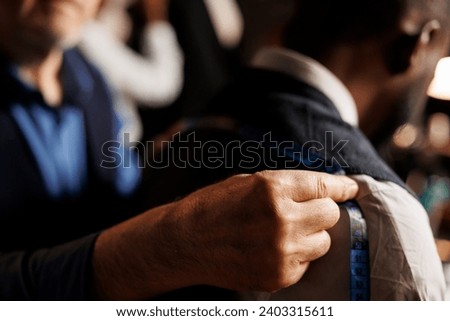 Close up of professional experienced suitmaker doing fitting length estimations on sartorial piece using measurement tape. Meticulous couturier helping client with custom handmade elegant suit
