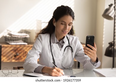 Close Up Professional Female Doctor Wearing Earphones Holding Phone, Consulting Patient Online, Taking Notes, Filling Medical Documents, Physician Therapist Making Video Call, Telemedicine Concept