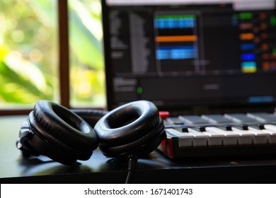 Close Up Of Professional Headphones And A Midi Keyboard In A Music Producer Home Studio. Laptop And Window With Nature In The Background.