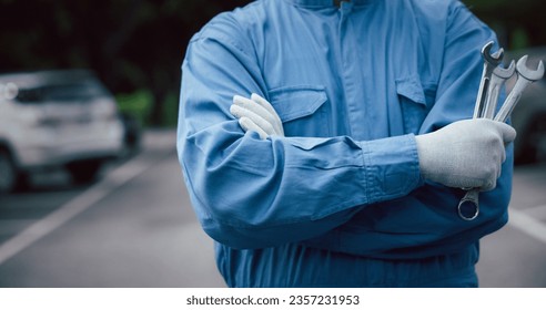 Close up of professional mechanic arm cross while holding wrench and wear blue uniform. Mechanic holding a spanner getting ready to work on a car at his workshop. Car mechanic proudly posing.