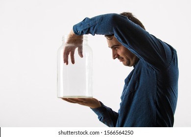 Close Up Profile Portrait Of A Curious Businessman Holding An Empty Glass Jar Try To Put His Hand Inside Looking With Sneaky Eyes Isolated Over White Background.