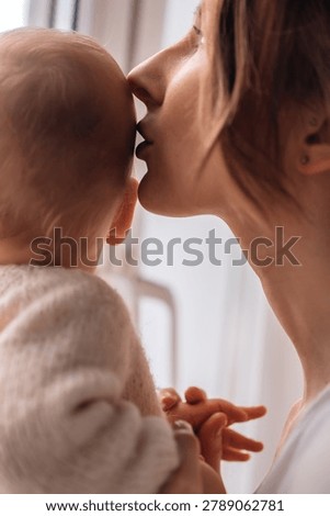 Close up profile view of a caring young mom gently kissing her small child forehead, holding tiny hands together in front of a bright window