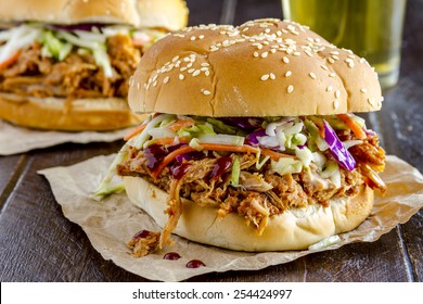 Close up of pulled pork barbeque sandwich with coleslaw sitting on wooden table with glass of beer