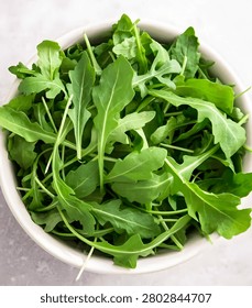 Close up of raw organic arugula or rocket salad leaves piled up in a ceramic white bowl over bright concrete background overhead view