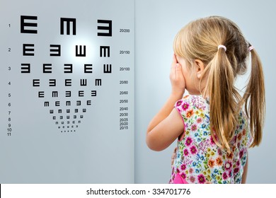 Close Up Rear View Of Little Girl Looking At Eye Test Chart.Kid Closing One Eye With Hand.