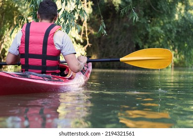 Close Up Of Rear View Of Man Kayaking On Red Kayak In The Summer River Near Green Trees