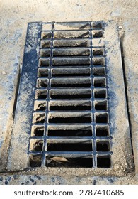 Close up of a rectangular metal drainage grate cover on a sandy, dusty ground.