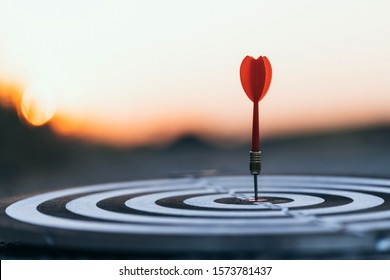 Close up red dart arrow hitting target center dartboard on sunset background. Business targeting and focus concept.