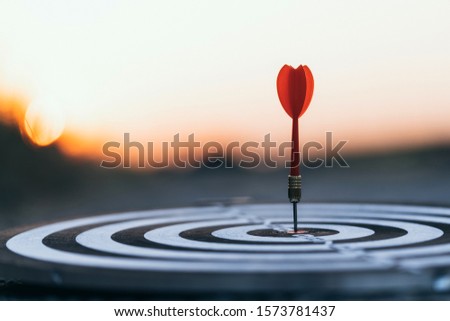 Close up red dart arrow hitting target center dartboard on sunset background. Business targeting and focus concept.