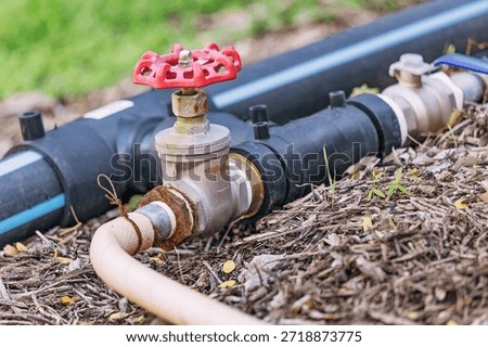 Close up of a red valve and rusty pipe connecting to a black main line, delivering water for outdoor irrigation