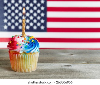 Close Up Of A Red, White, And Blue Frosted Cupcake With Burning Candle On Rustic Wood. Fourth Of July Concept With United States Of American Flag In Background.