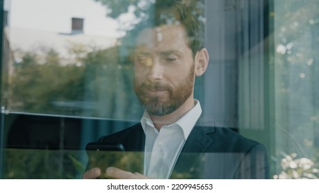 Close Up Reflection In Glassy Window Blue Glass Caucasian Bearded 40s Middle-aged Man Adult Businessman Walking Indoor In Office Company Browsing Scrolling Phone Mobile App Looking Outside Pensive