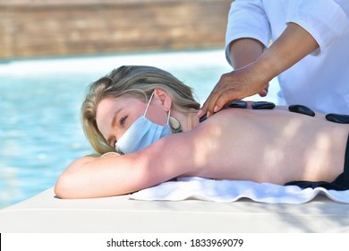 Close Up Of A Relaxed Looking Blonde Woman Wearing A Surgical Mask Enjoying A Hot Stone Massage On An Out Of Focus Background. Safety And Well-being .