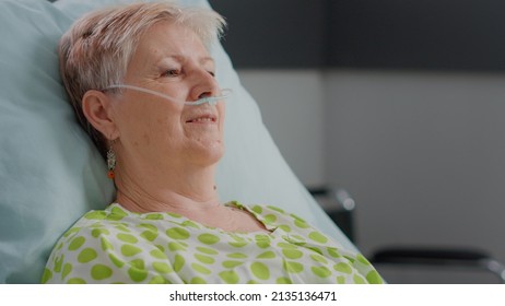 Close Up Of Retired Patient With Illness Having Nasal Oxygen Tube And Sitting In Hospital Ward Bed. Senior Woman Preparing To Be Treated Against Disease, Hoping For Recovery At Clinic.