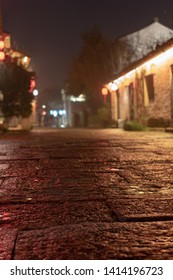 Close Up Rock Paved Road Of Chinese Ancient Town In The Night. Rough Surface With Rain Water. Defocused Ancient Building And Tourists Background. Low Angle