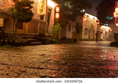 Close Up Rock Paved Road Of Chinese Ancient Town In The Night. Rough Surface With Rain Water. Defocused Ancient Building And Tourists Background. Low Angle