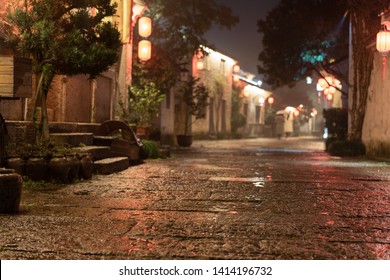 Close Up Rock Paved Road Of Chinese Ancient Town In The Night. Rough Surface With Rain Water. Defocused Ancient Building And Tourists Background. Low Angle