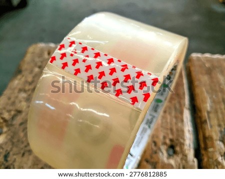 Close up of a roll of clear adhesive packaging tape with red arrow markings on a wooden surface. Professional industrial stationery for shipping and logistics.