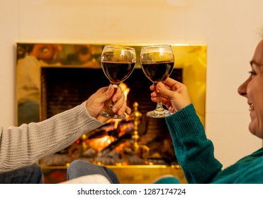 Close Up Of Romantic Couple Holding Two Red Wine Glasses Toasting At Fire Celebrating Happy Moment Together By Fireplace At Cabin Home In Winter Holiday Vacation, Christmas Or Anniversary Concept.