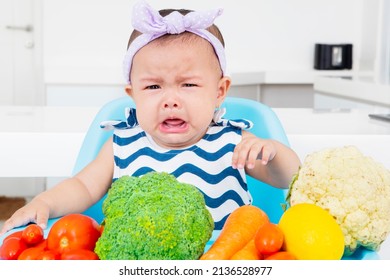 Close Up Of Sad Baby Girl Making A Pout Face While Sitting On The Baby Chair With Fresh Vegetables In The Kitchen. Shot At Home