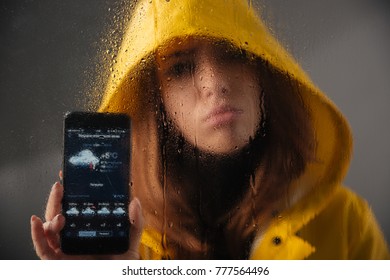 Close Up Of A Sad Girl Dressed In Raincoat With A Hood On Her Head Standing Behind A Wet With Rain Window And Showing Mobile Phone With A Weather App On A Screen Isolated Over Gray Background