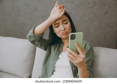Close up sad young woman of wears casual clothes hold in hand use mobile cell phone put hand head sits on grey sofa couch stay at home hotel flat rest relax spend free spare time in living room indoor