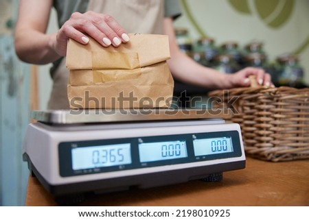 Close Up Of Sales Assistant In Sustainable Plastic Free Grocery Store Weighing Goods In Paper Bag On Digital Scales