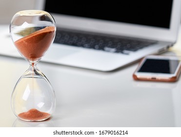 Close up of sandglass with laptop and phone. Close up of home office workspace, desktop. Copyspace for text.