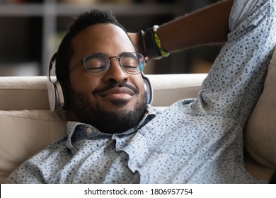Close up satisfied African American man wearing headphones enjoying favorite music with closed eyes, daydreaming or sleeping, positive young male wearing glasses lying relaxing on cozy couch