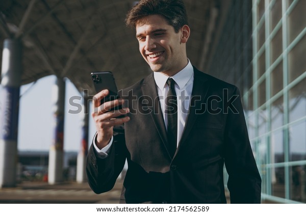 Close up satisfied young traveler businessman\
man in black suit standing outside at international airport\
terminal use mobile cell phone book taxi order hotel online Air\
flight business trip\
concept.