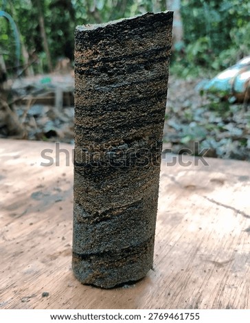 Close up of sedimentary rock core sample displying layered structure with dark carbon-rich bands, posibly indicating organic material or coal presence. Geological specimen photograped in outdoor setting 
