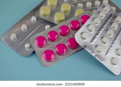 Close up selective focus of assorted prescription pills in silver foil blister packs on a baby blue background. Different types of colored tablets in packets piled and cropped on a sky blue backdrop.