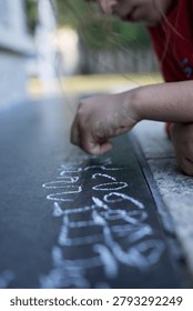 Close up selective focus on a sequence of hand written white chalk numbers on a dark stone tile pavement with a blurred kid in red shirt in the background. Concept of early education, math practice and outdoor school activities.