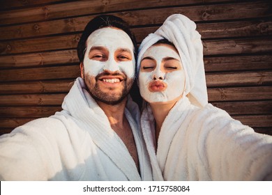 Close up selfie of beautiful happy couple after shower with a towel on head sending air kiss with cream mask on face.Woman with clay mask taking selfie with mobile phone at home enjoying relaxation.