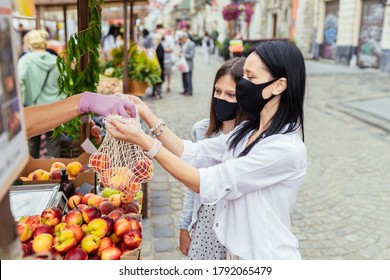 Close Up Of Seller's Hads In Protective Gloves Putting Organic Peach In Mesh Bag. Two Female Buyers In Protective Masks Chooses Peaches In Farmer Market. New Social Rules After Pandemic.