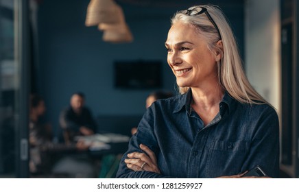 Close Up Of Senior Business Woman Standing In Office With Her Arms Crossed And Looking Away Smiling. Mature Female In Office With Team Meeting In Background.