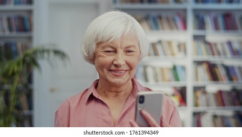 Close Up Of The Senior Grey-haired Pretty Smiled Woman Talking Via Videochat On The Smartphone Joyfully As Using Web Cam. At The Library.