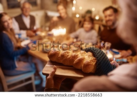 Close up of senior Jewish woman bringing Challah bread at dining table while having meal with her extended family on Hanukkah.