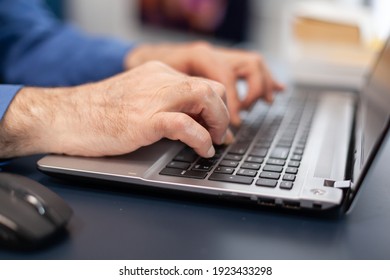 Close Up Of Senior Man Hands Typing On Laptop Keyboard. Elderly Man Entrepreneur In Home Workplace Using Portable Computer Sitting At Desk While Wife Is Holding Tv Remote