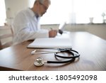 Close up senior mature doctor work desk with stethoscope, middle aged therapist practitioner gp sitting at table, writing in documents, filling illness history, patient card, working in hospital